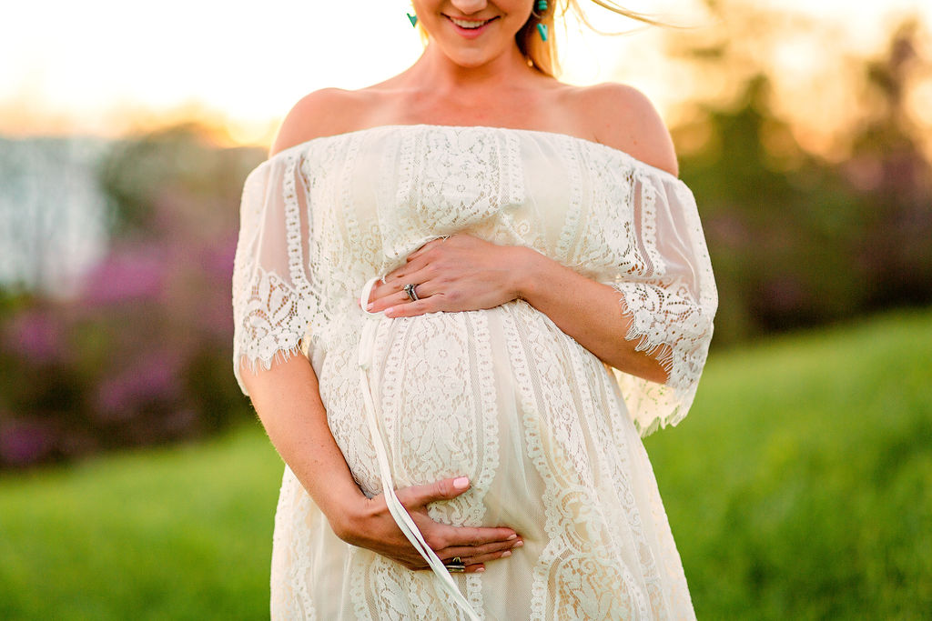 woman wearing white maternity gown stands in a field holding her bump