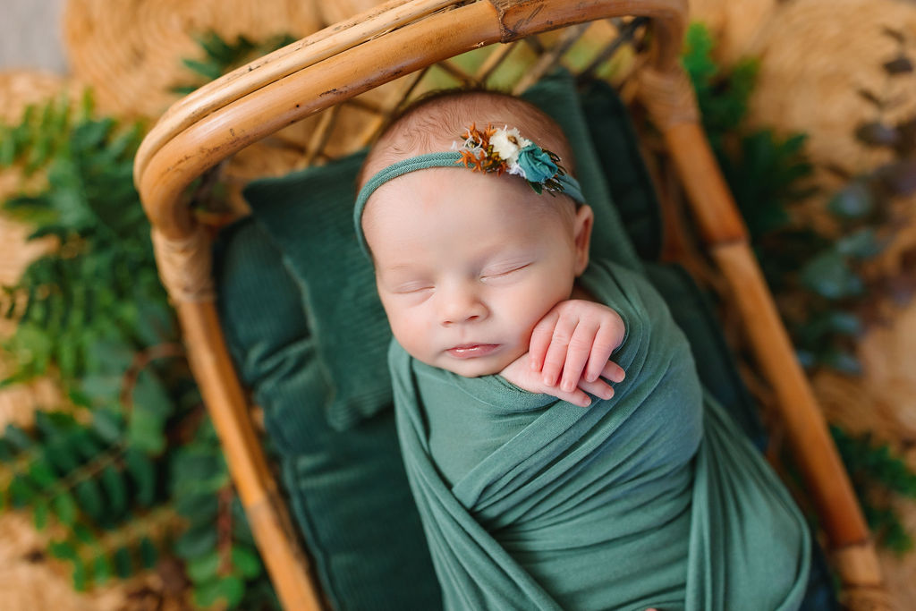newborn in a green swaddle and headband sleeps with hands out