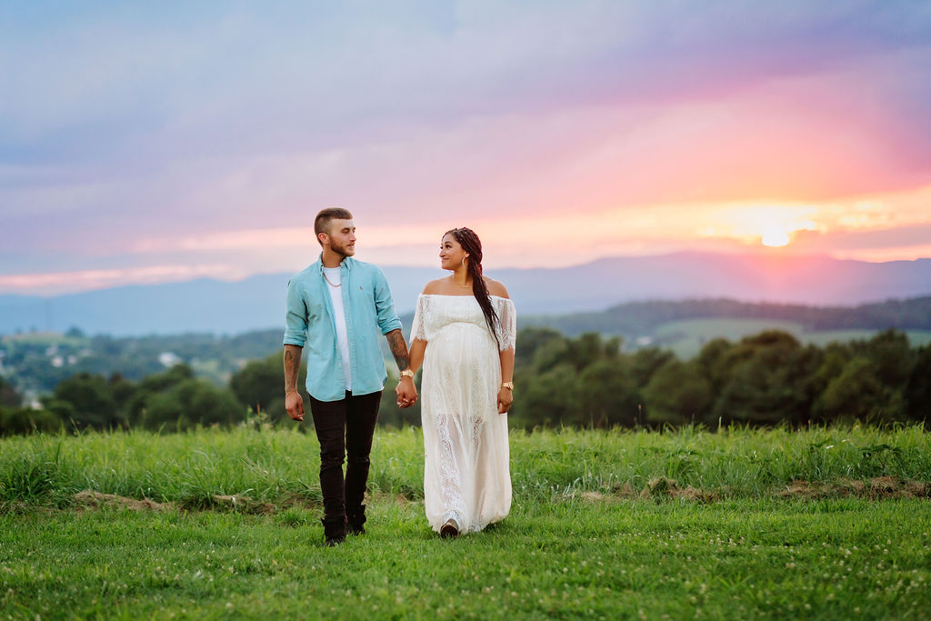 mother to be wearing a white maternity gown walks in a grassy field holding hands with husband