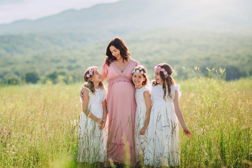 Mother to be stands in tall grass with mountains iin the background with her three older daughters prenatal yoga charlottesville