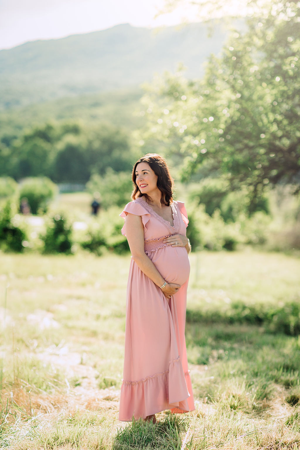 Mother to be wearing a pink maternity gown stands in a grassy field by a tree prenatal yoga charlottesville