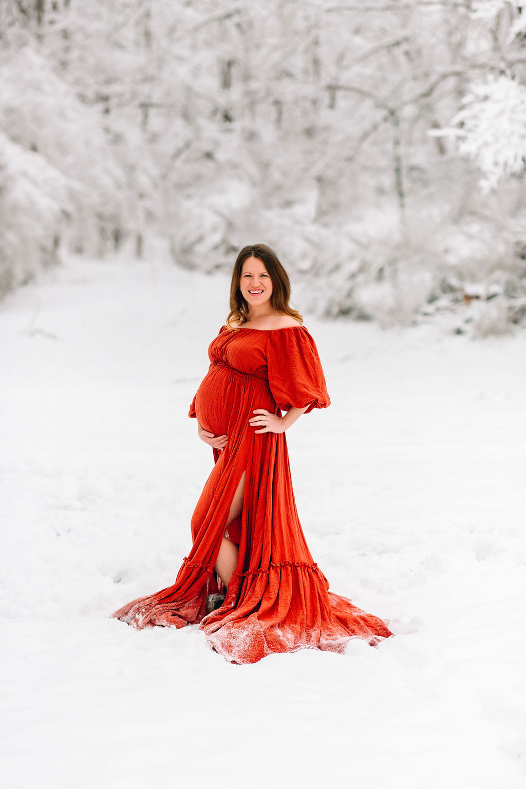 Mother to be wearing a long red maternity gown stands in a snowy forest uva midwives