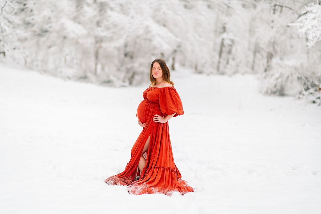 mother to be wearing a red maternity gown stands in fresh snow holding her bump uva midwives