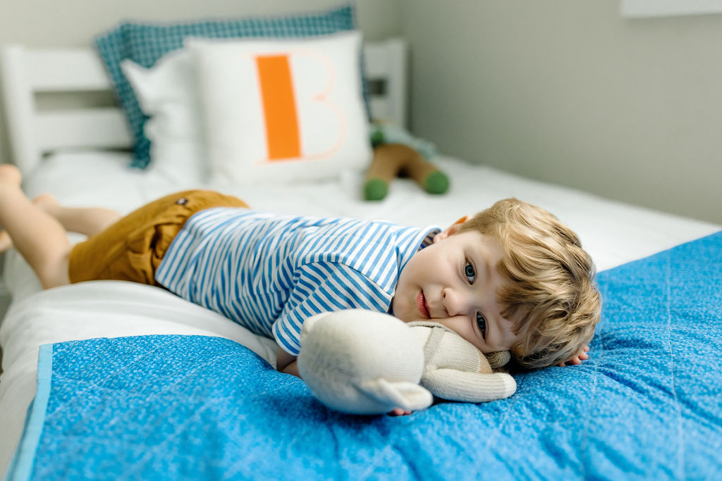boy wearing shorts and blue polo lays on his bed cuddling with knit stuffed toy