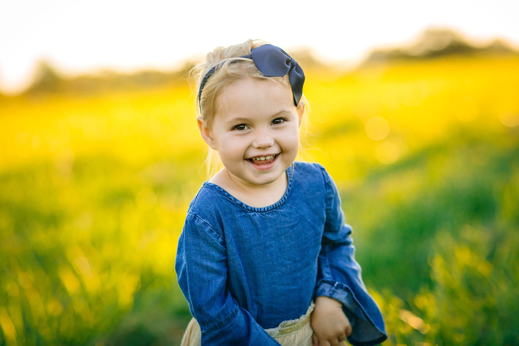 Young toddler girl plays in a grassy field wearing a blue bow and denim top
