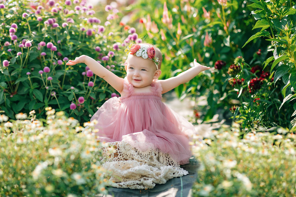A toddler girl throws her arms in the air while playing in a flower garden in a pink velour dress ashby mae boutique