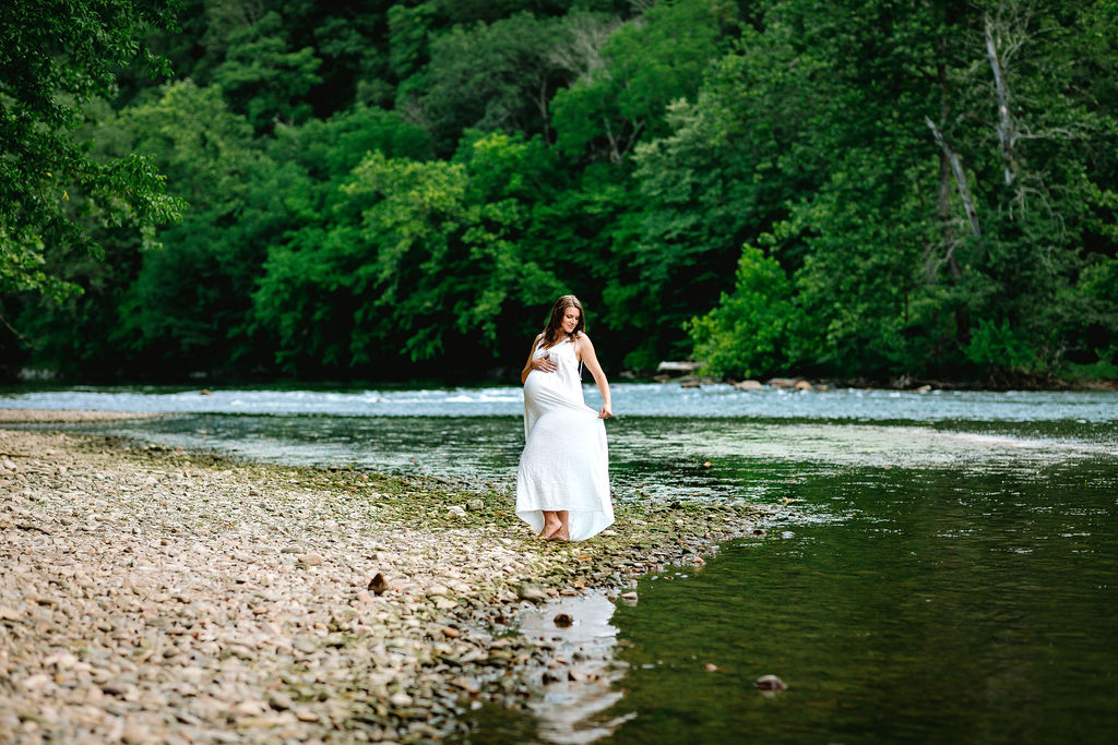 A mom to be holds her white dress up as she walks along a rocky river's edge in a forest