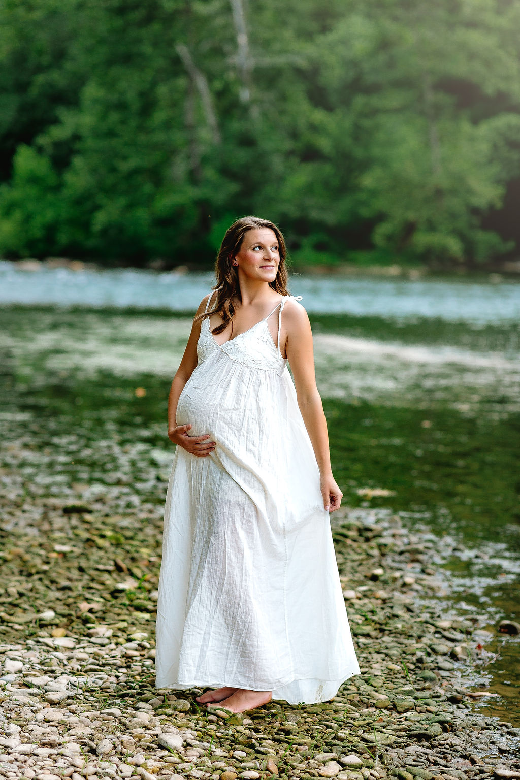 A pregnant woman stands on a rocky riverbed in a white maternity gown while holding her dress and bump belle grove birth center