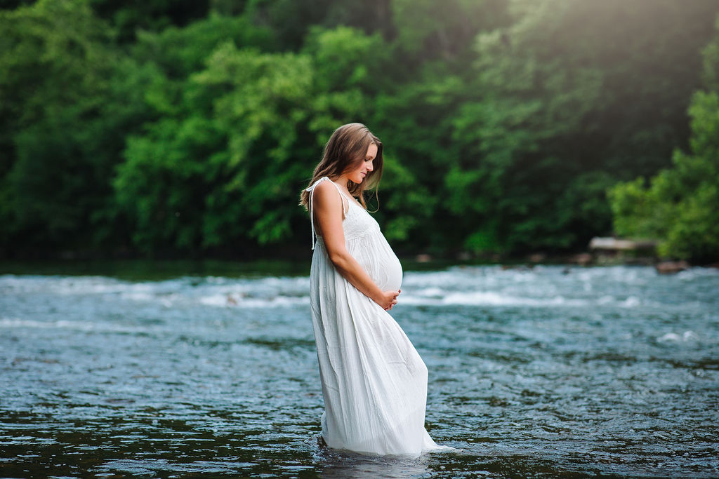 A mother to be in a white maternity gown stands in a flowing shallow river while holding her bump belle grove birth center