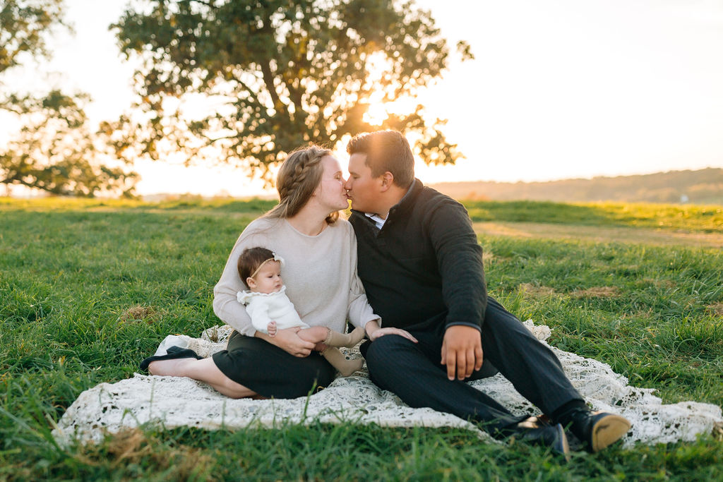 New parents kiss while sitting on a white blanket in a grassy field holding a newborn baby charlottesville pediatricians