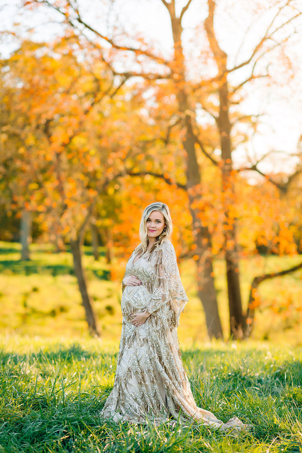 A mother to be in a long ornate maternity gown holds her bump while standing in a grassy green field with fall colored trees behind her at sunset obgyn winchester va