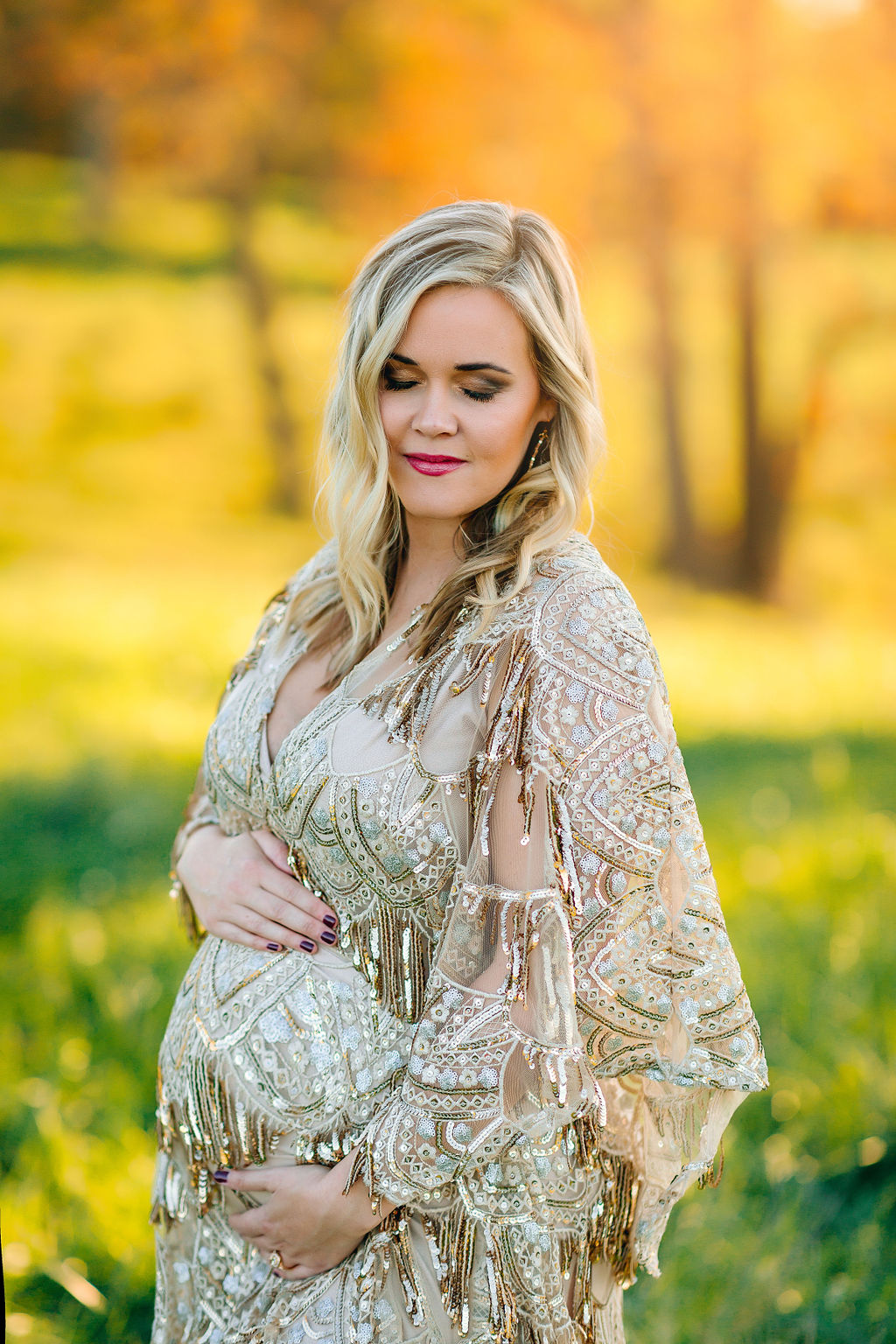 A mom-to-be wears an ornate maternity gown while standing in a green field with golden trees behind her obgyn winchester va