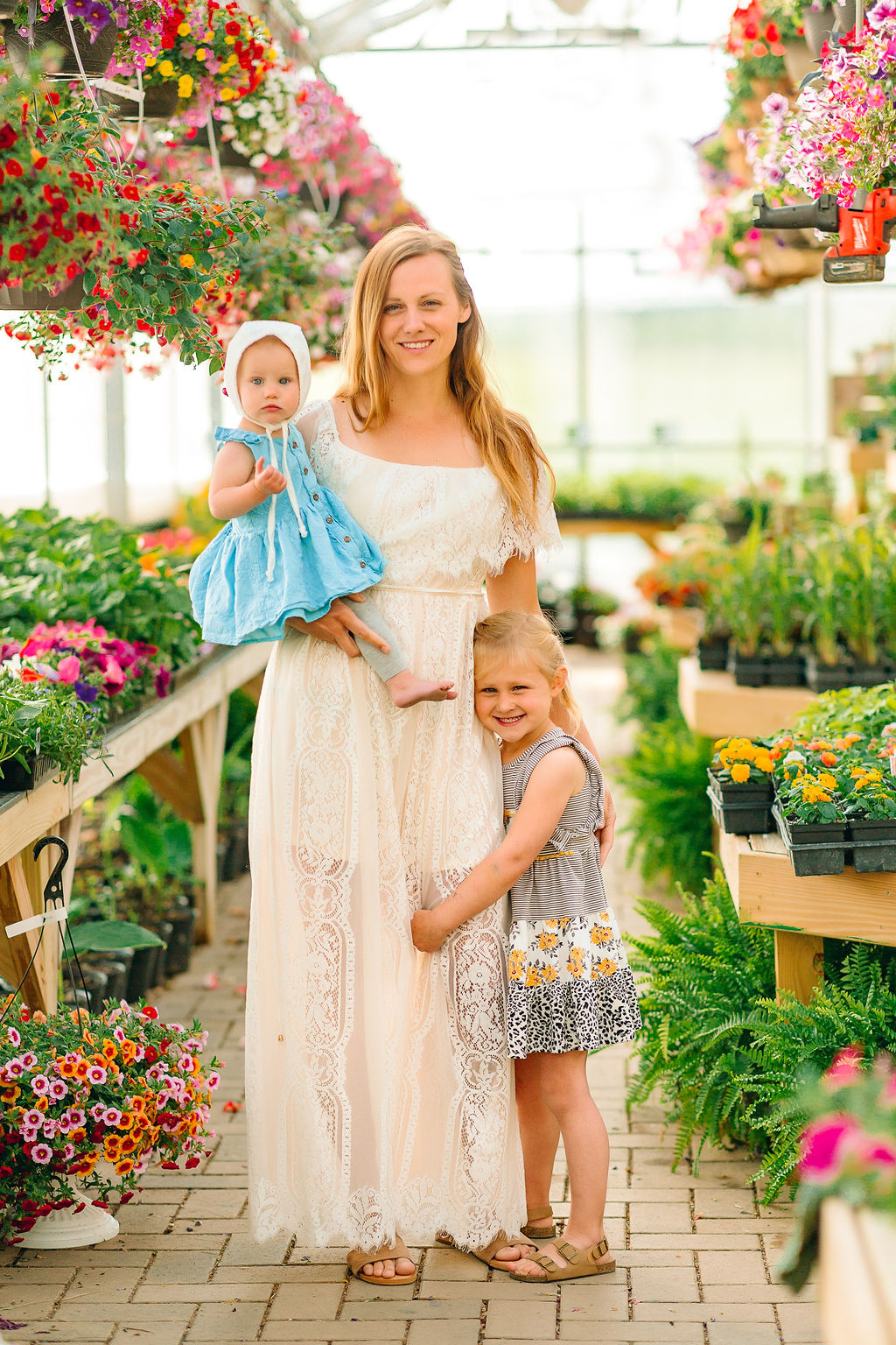 A mother stands in a flower filled greenhouse with her toddler daughter and older daughter
