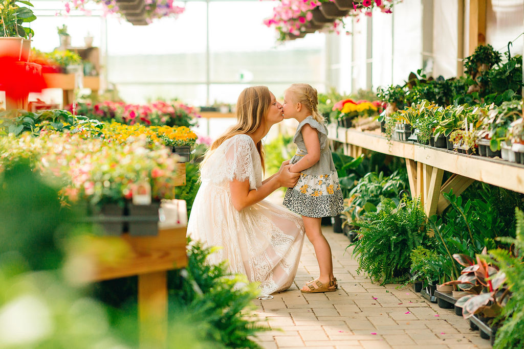 A mother kisses her daughter in an aisle of a greenhouse full of flowers pediatricians in winchester va