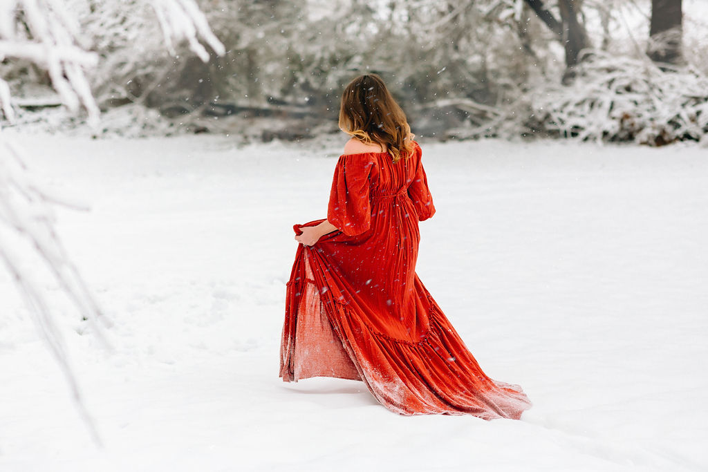 A mother to be carries her red maternity dress while walking through deep snow