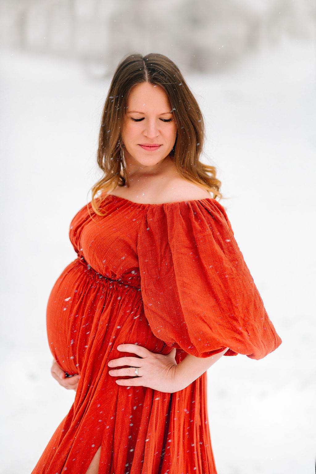 A mother to be in a red maternity dress stands in snow looking down her shoulder premier birth center