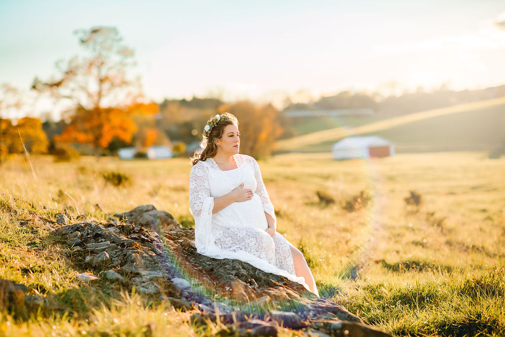 Mother to be sits on some rocks in a field in a white maternity gown