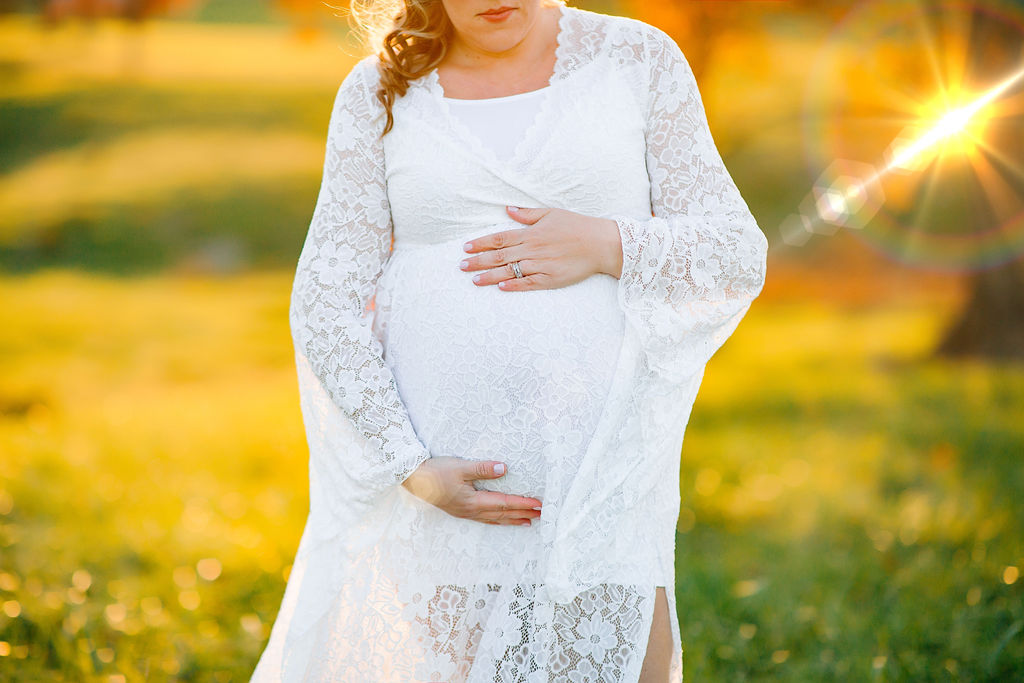 Mother to be stands in a golden grassy field in a white lace maternity gown prenatal massage charlottesville