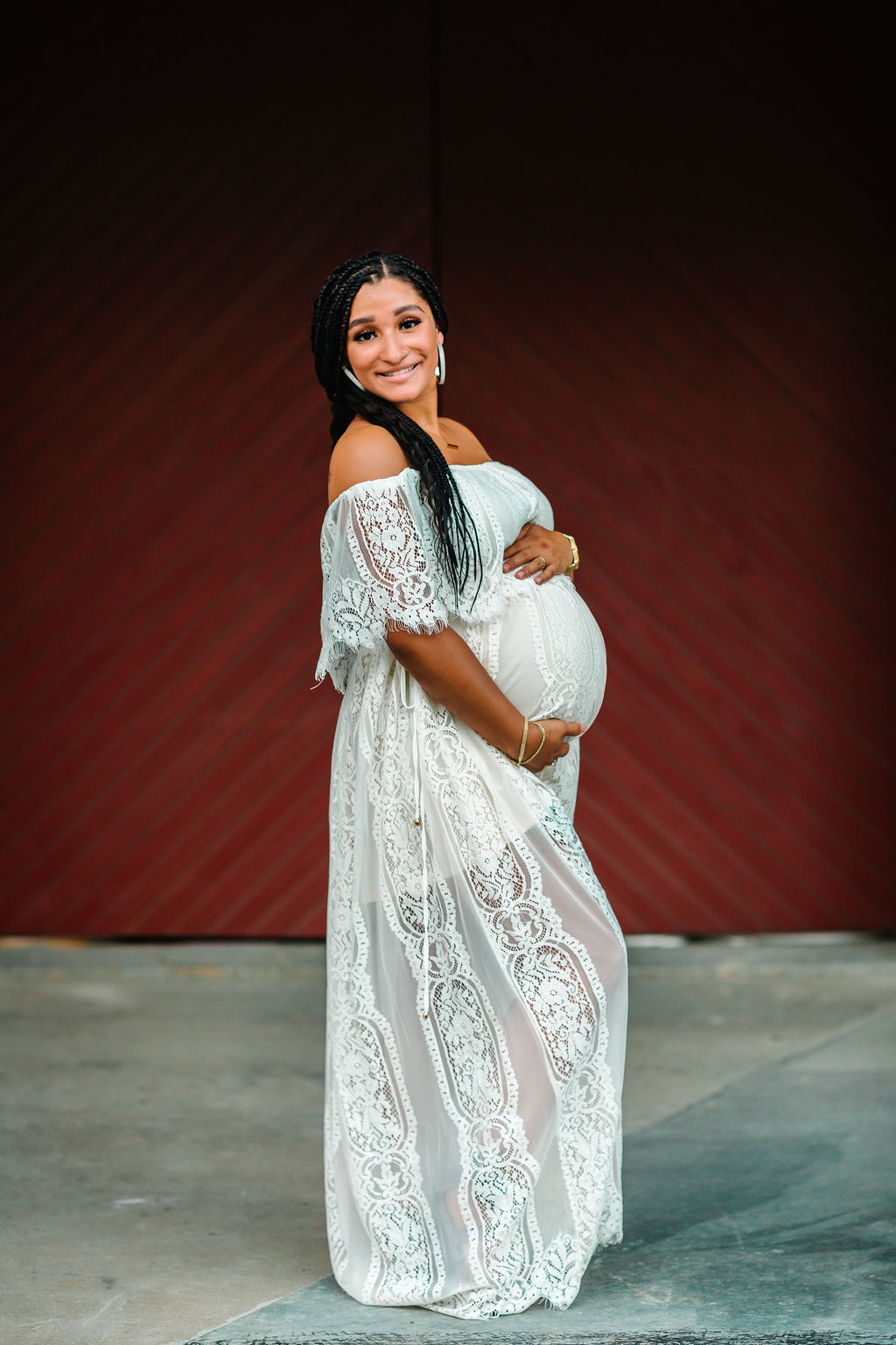 Mother to be stands in front of a red wall wearing a white maternity dress