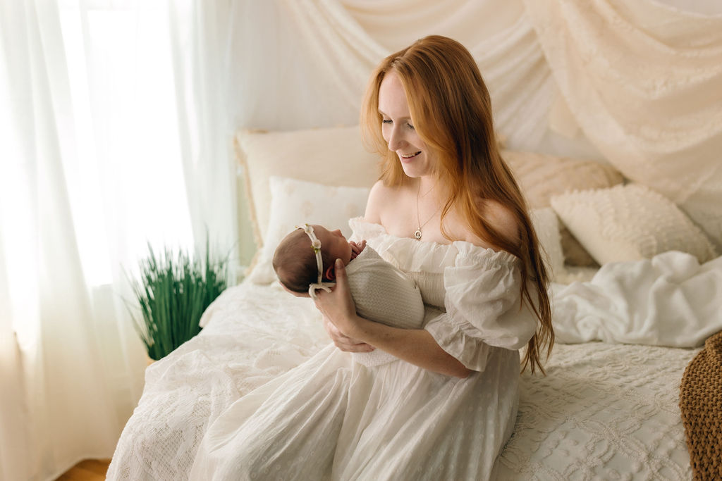 A mother sits on the edge of a bed wearing a white flowy dress while looking down at her newborn baby in her hands