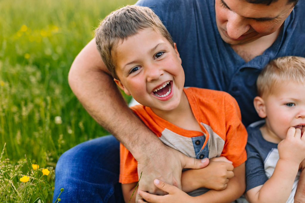 A young boy sits in his dad's lap while being tickled in a grassy field with wildflowers