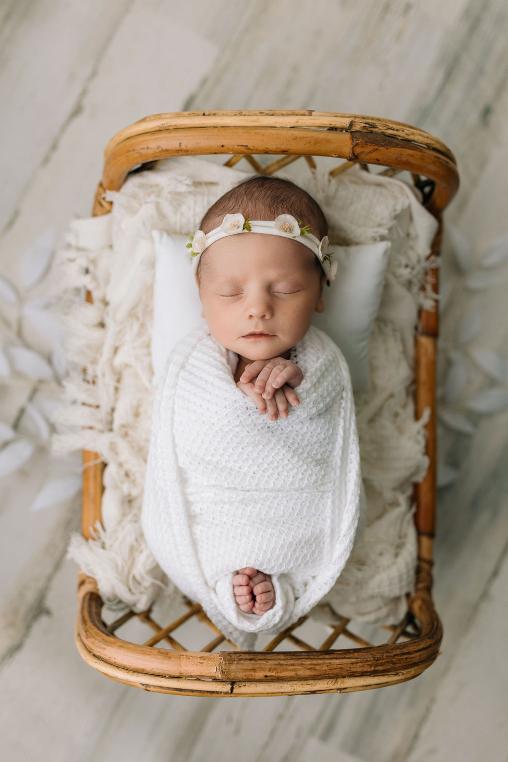 A newborn baby sleeps in a white swaddle with toes and fingers sticking out and a white flower headband in a tiny wicker bed daycare staunton va