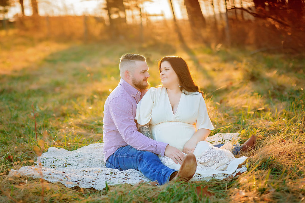 A mother to be in a white maternity dress sits on a lace blanket in a grassy field with her husband at her side grace midwifery