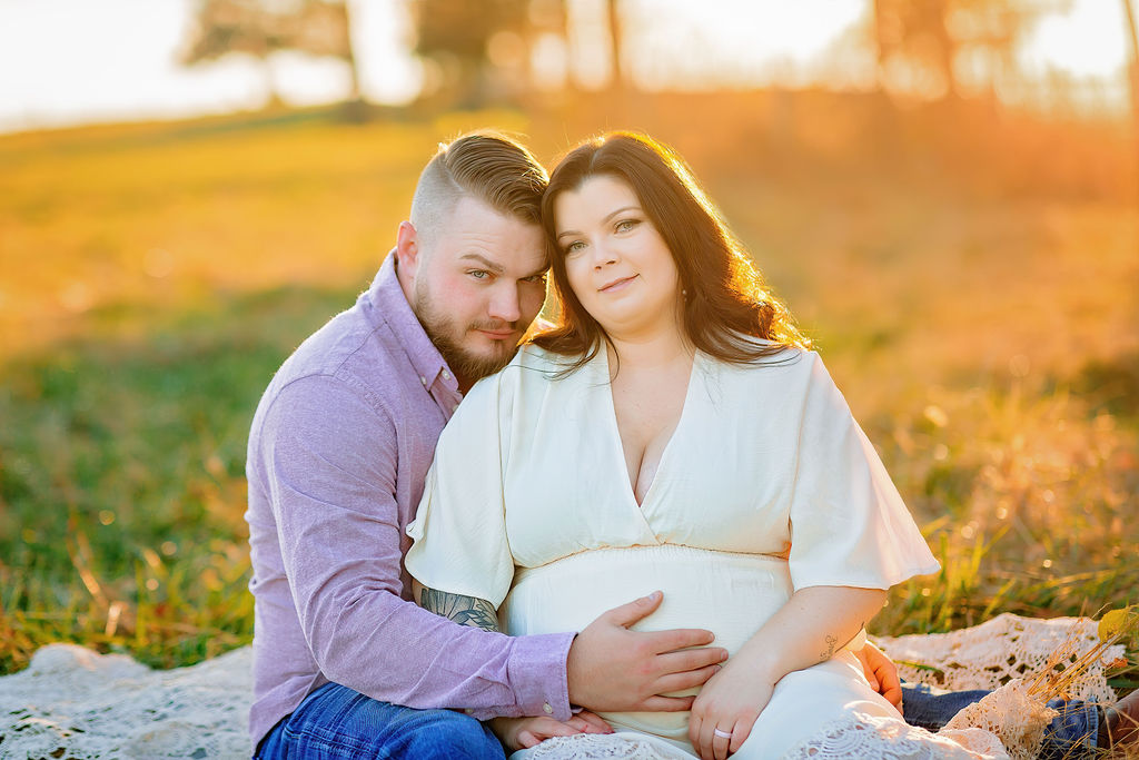 A mother to be in a white maternity dress sits with her husband in a field on a lace blanket
