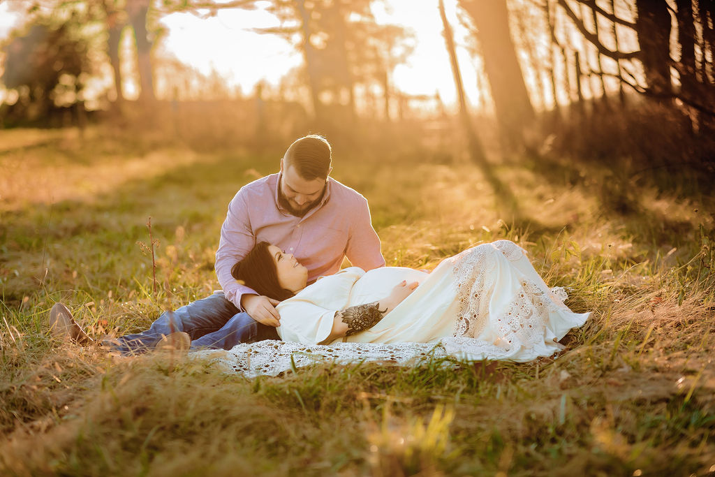 A mother to be in a white lace maternity dress lays her head on the lap of her husband in a grassy tree lined field at sunset grace midwifery