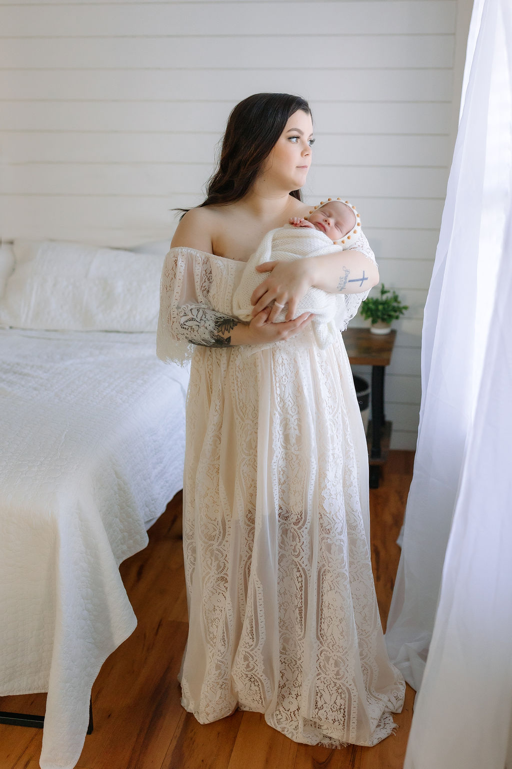 A new mom holds her sleeping newborn daughter in a white lace dress while looking out a window lactation consultant winchester va