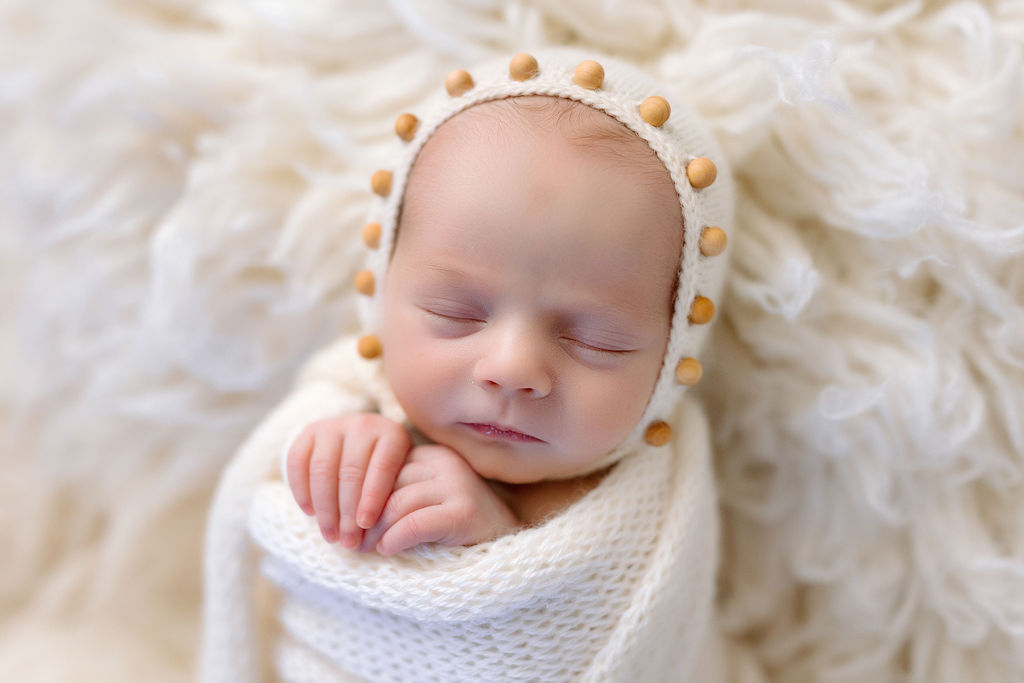 A newborn baby sleeps while swaddled in a knit white blanket with hands poking out and wearing a white knit hat with small wooden balls lining the edge