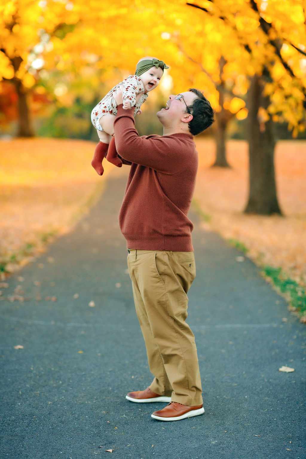 A dad and toddler daughter play in the fall on a paved park path
