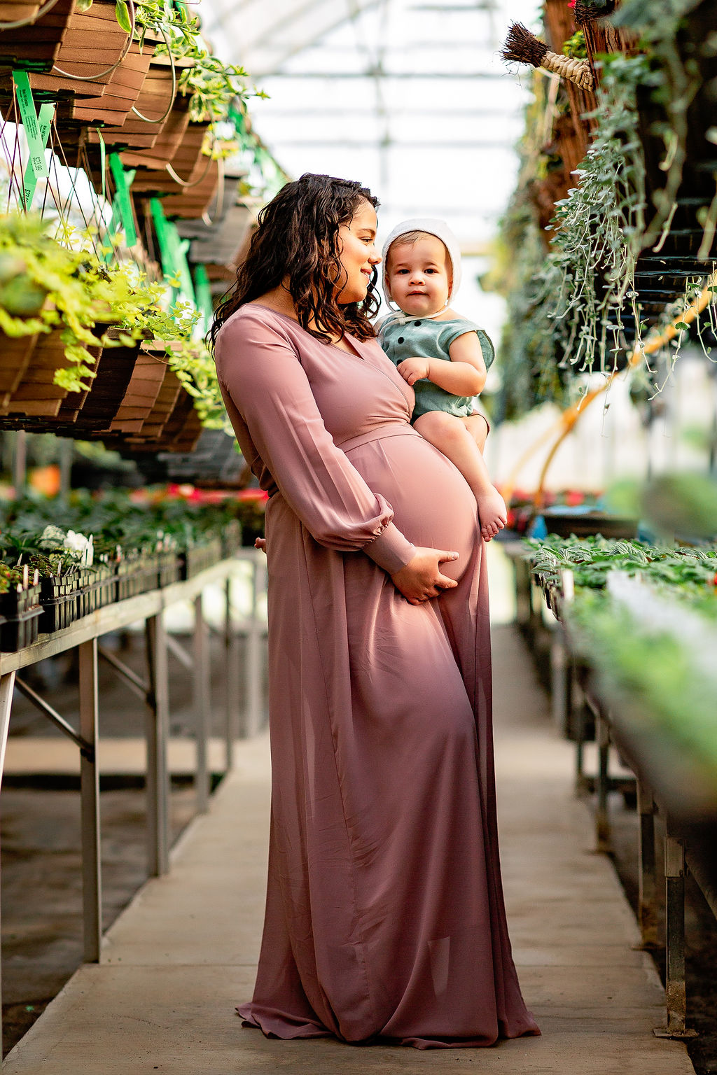 A mother to be browses a greenhouse aisle in a purple maternity gown while holding her toddler son marshall midwifery
