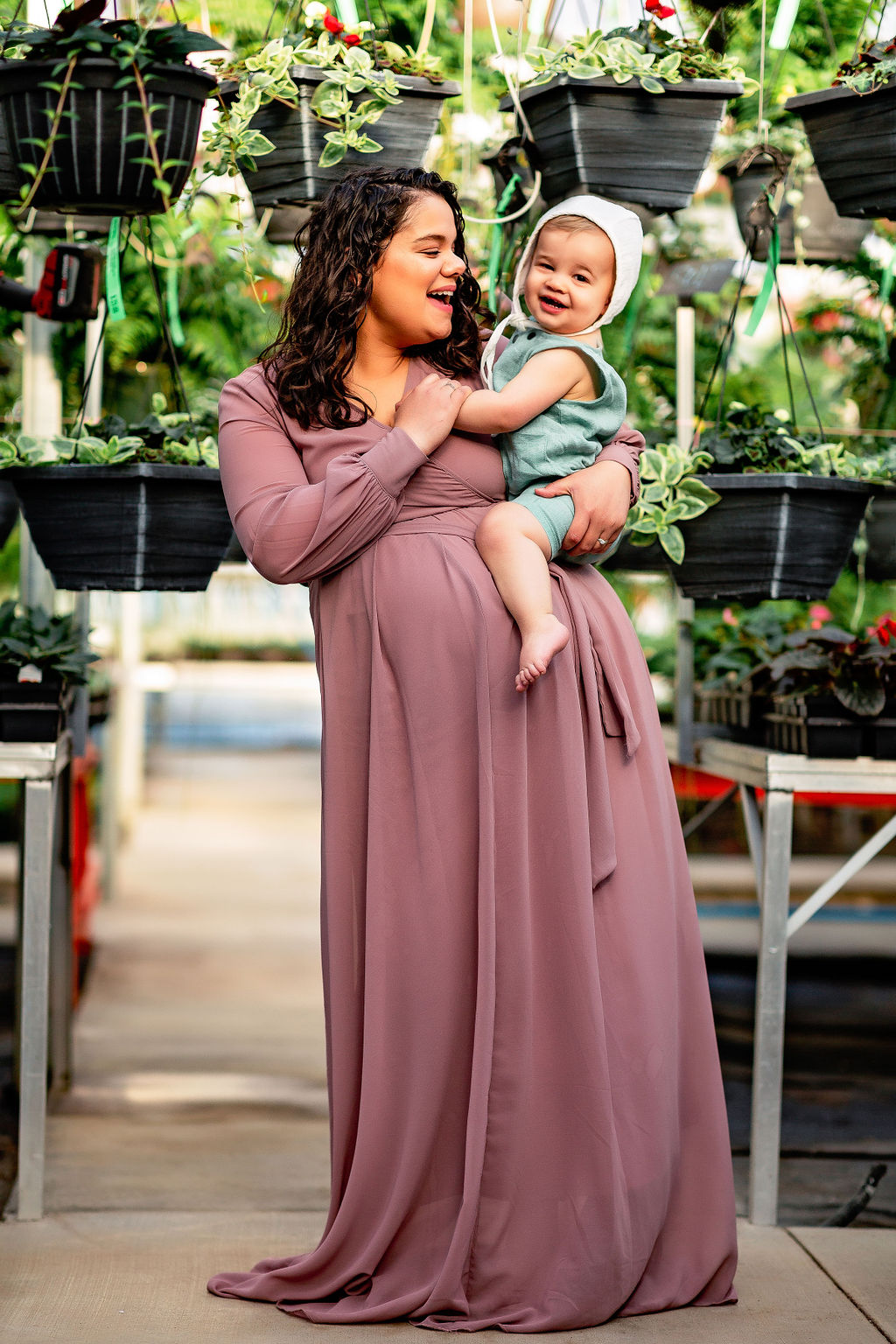 A pregnant woman stands in a greenhouse surrounded by hanging plants in a maroon maternity gown while holding her toddler on her hip marshall midwifery