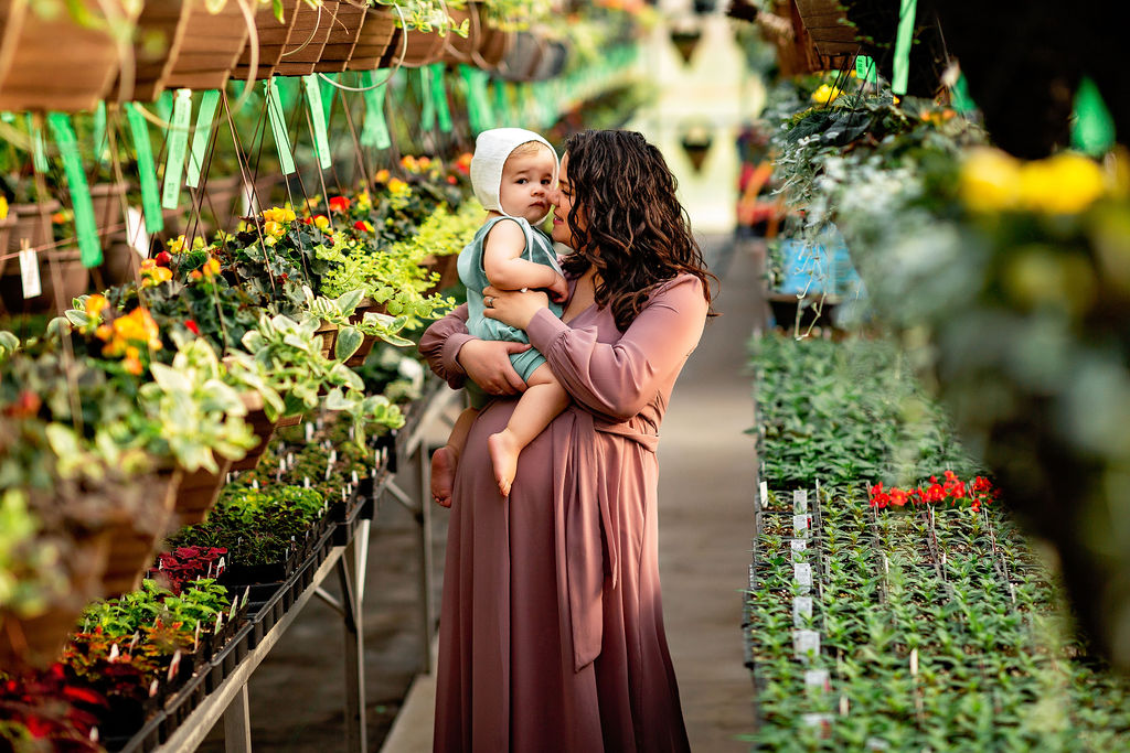 A pregnant mother plays with her toddler son iin an aisle of a greenhouse filled with plants