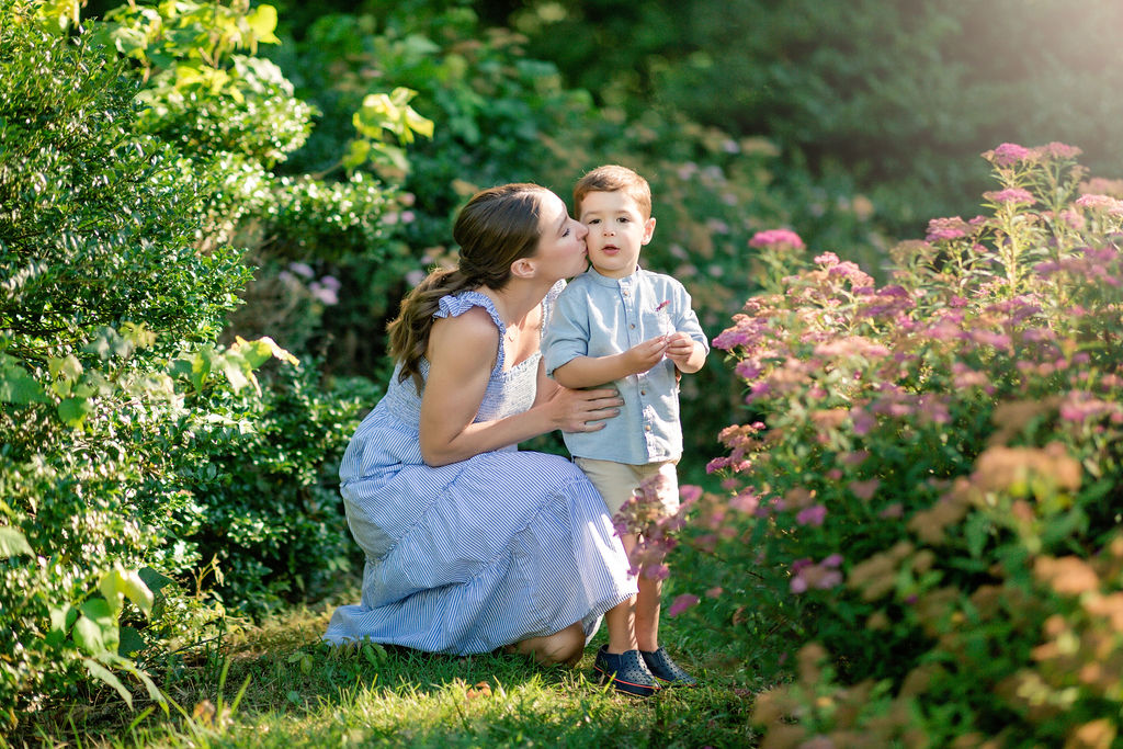 A mother kneeling in a flower gardeen path kisses the cheek of her son who is holding a pink flower mommy and me classes charlottesville va
