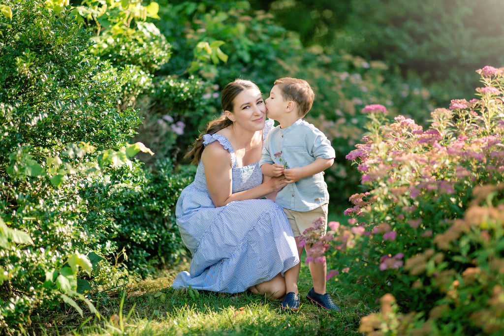 A mother kneels in a glower garden with her young son who is kissing her on the cheek in a blue dress mommy and me classes charlottesville va