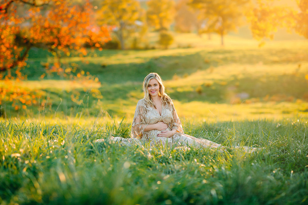 A mother to be in a tan maternity gown sits in a grassy field with her hands on her bump at sunset obgyn front royal va