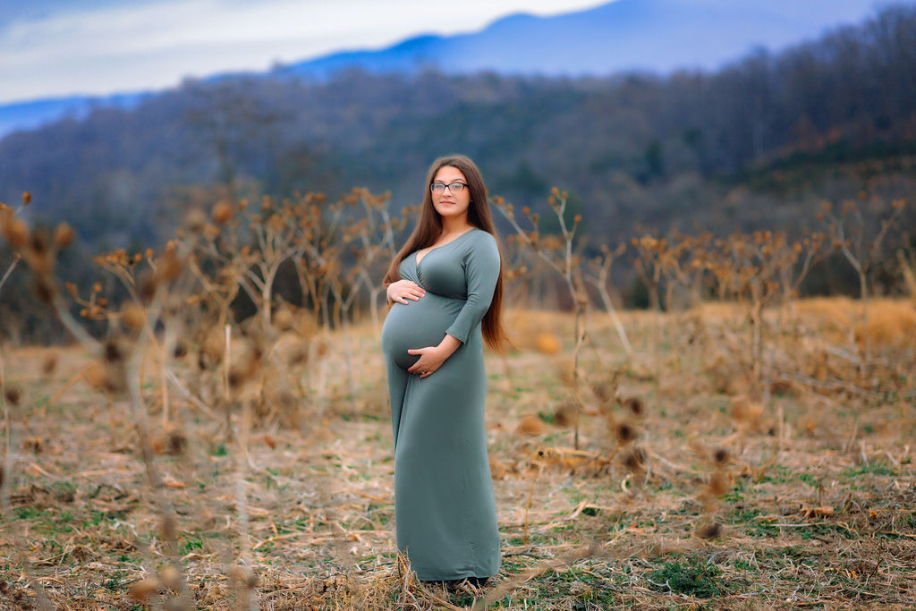 A mother to be stands in a field with the blue ridge mountains behind her in a blue maternity gown