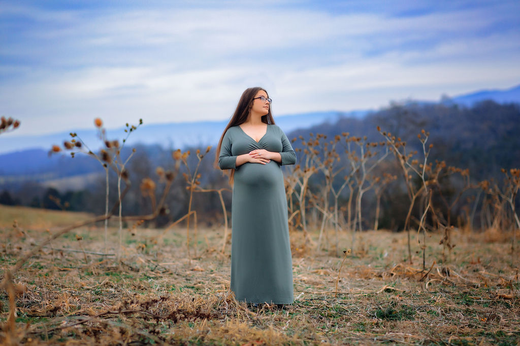 A mother to be stands in a field on a hill with the blue ridge mountains behind her in a blue maternity gown prenatal massage winchester va