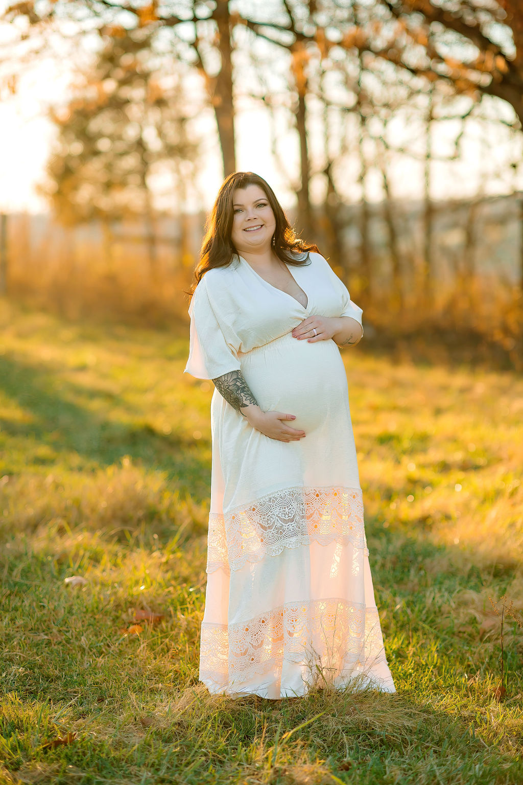 A mother to be holds her bump in a grassy field at sunset in a white maternity dress with a lace bottom prenatal yoga winchester va