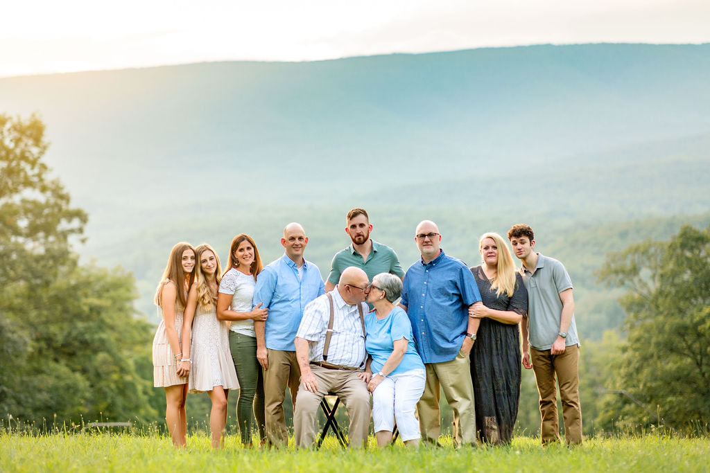 Grandma and Grandpa kiss while sitting on stools on a hillside while their family surrounds them