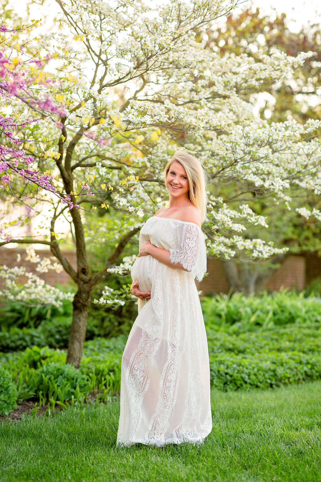 A mother to be stands in a garden with blooming dogwood trees in a white lace maternity gown