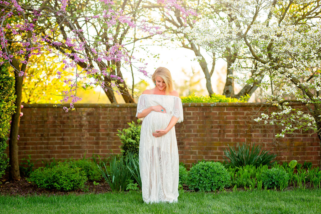 A mother to be in a long white maternity dress stands in front of a brick wall under blooming trees looking down at her bump augusta healthcare for women