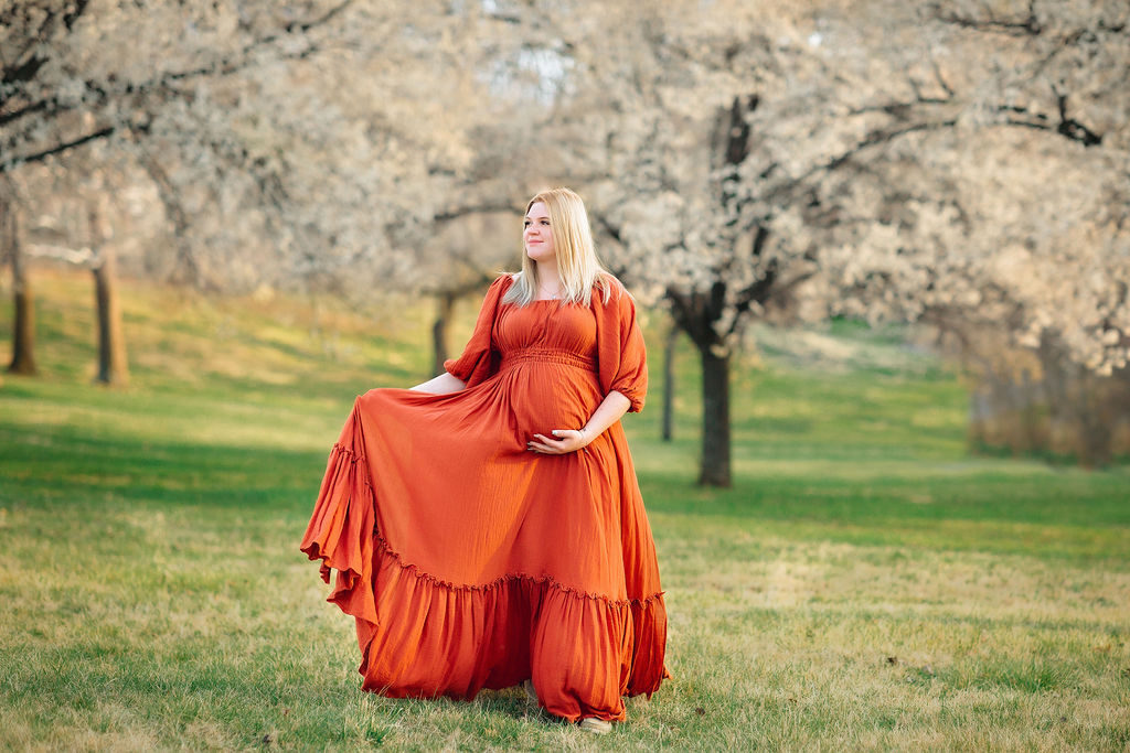 A mother to be in a red maternity dress walks through a field of flowering white trees