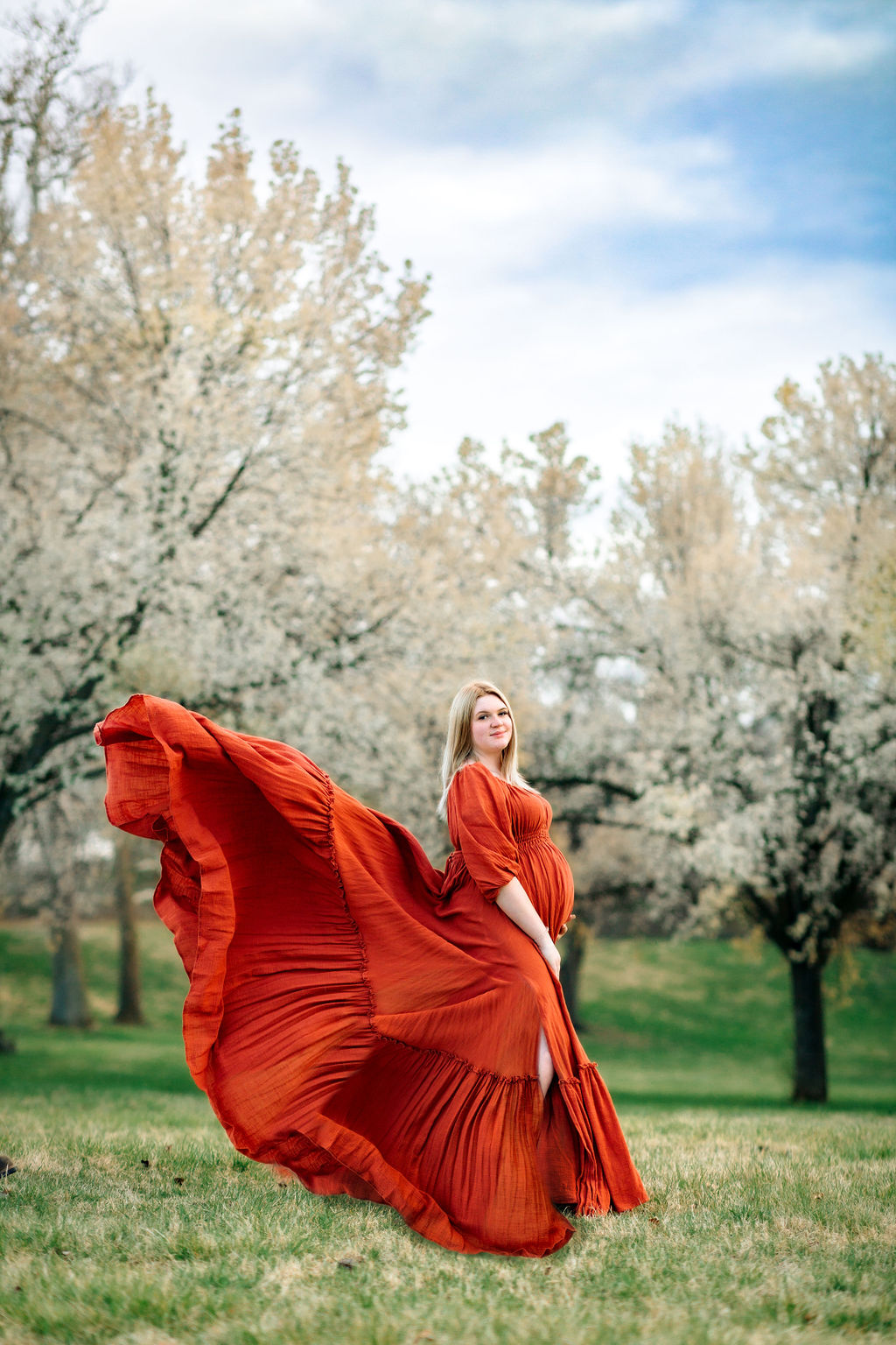 A mom to be stands among a group of flowering trees in a bright red maternity gown that is flowing in thee wind behind her babymoon destinations virginia