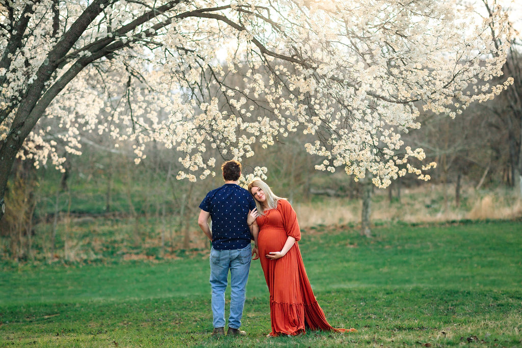 A mother-to-be in a bright red dress leans onto the shoulder of her partner while standing in a field under a flowering tree babymoon destinations virginia