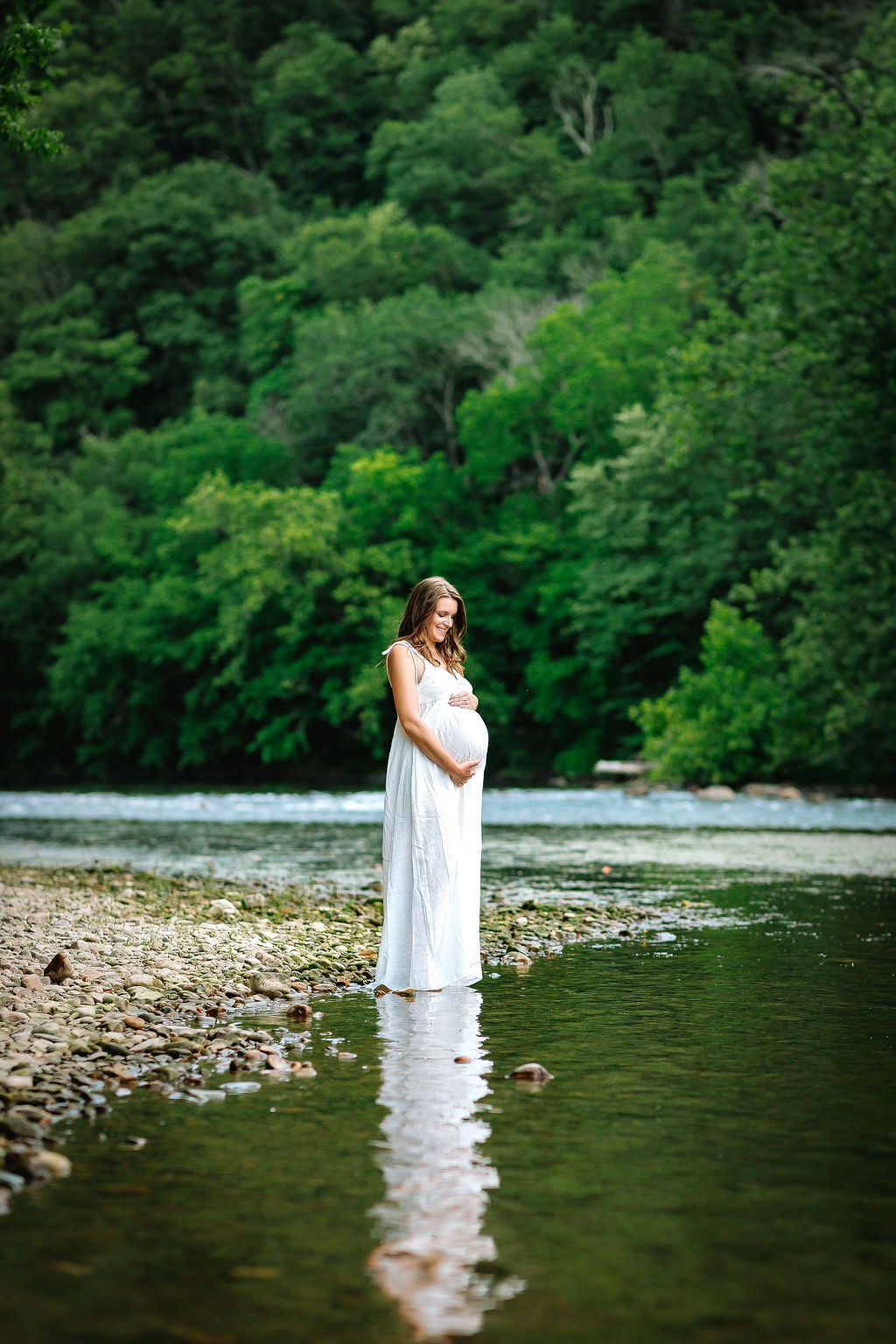 A mother to be stands on a river's edge looking down at her bump in a long white maternity dress jefferson obgyn