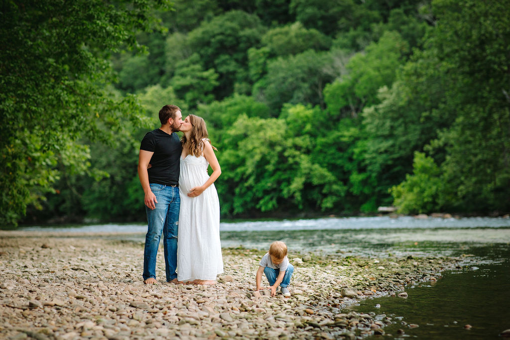 A pregnant couple kisses while their son plays with rocks next to a river