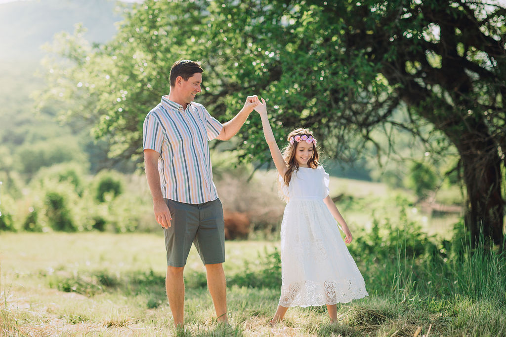 A father dances with his daughter in a white lace dress under a tree moutain ridge pediatrics